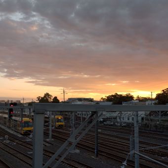 Sunset Above The Trainyards, Hornsby, with grey, fluffy clouds lined with streaks of gold eminating from the centre right, with two parked trains in the bottom left corner on tracks which lead to the bottom right corner