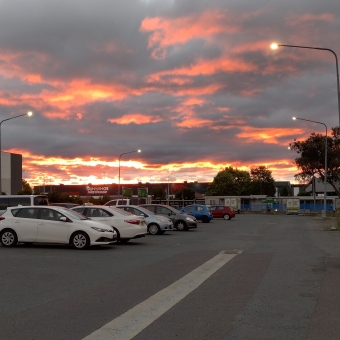 A Fire-Like Sunset, Gungahlin, with a red hot glow from the setting sun on the horizon barely visible above a shopping centre, with a carpark filled with streetlights and different-coloured cars in the foreground