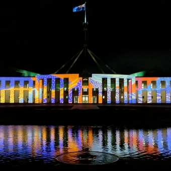 Reconciliation Lights, New Parliament House, with New Parliament House from and centre seemingly covered in Indigenous art which is a mosaic of coloured pattertned dots and dashes which has some sections of blue and some of yellow and some of orange and one of purple and one of pink and one of green, with the Australian flag visible atop its mast, with the sky behind pitch black, with the reflection of all light in the shallow pool of water below