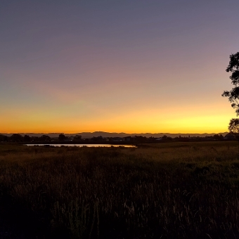 Radiating Sunset, Little Mulligans, Bonner, with a glowing orange sun strewn across a cloudless sky changing from gold to orange to magenta and finally fading to blue, with grass turning into low rolling hills silhouetted below