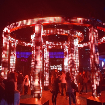 VIVID on Fire, Darling Harbour, with the word VIVID outlined by fire like a la neon signage visible through the stands of a pagola where every surface is covered in red LED lights