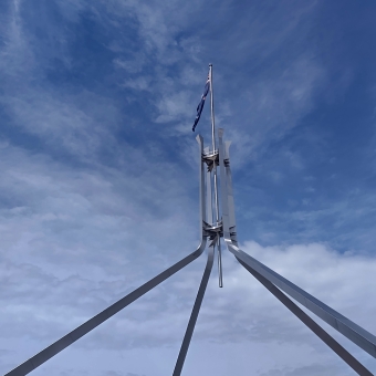 Blue Spire, Parliament House, Canberra, with the four large legs of the mast reaching from the bottom of the frame which levitate the single flag mast up amongst a brilliant blue with faint fluffy clouds