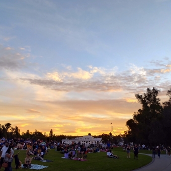 Parliamentary Sunset, Commonwealth Place, Parkes, with a streaked orange sunset over a wide crowd of people, with the silhouette of Old Parliament House and New Parliament House against the sunset in the distance