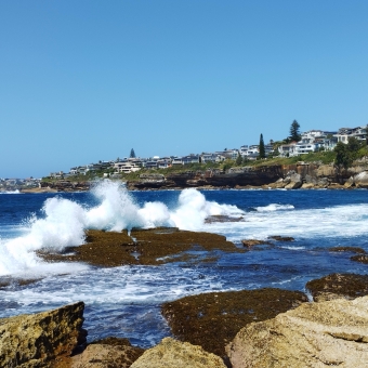 Wedding Cake Lookout, South Coogee, with a wave splashing against the rocks in the foreground, with the coast stretching from right to centre-left and the ocean to the left