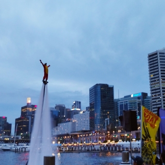 Lunar New Year, Darling Harbour, with someone in a dragon costume on a water-jetpack over the water and crowds in front of him on land and all against the backdrop of Darling Harbour high-rises