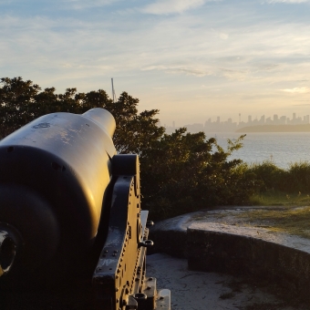 Historic Cannon, Camp Cove, Watsons Bay, with a cannon on the left against a backdrop of bushes, with Sydney city skyline silhouetted amidst a golden glow in the distance across vast water