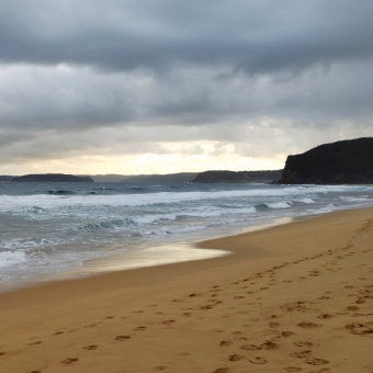Golden Views, Killcare Beach, Killcare, with golden sands covered in footprints and whitewashed waves against darkened headlands underneath grey clouds with a golden glow of sunset emanating from the horizon