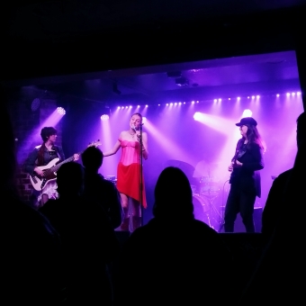 Gia Darcy at the Pop Up Pop Party, The Landsdowne Hotel, Chippendale, with singer Gia Darcy standing centre-stage in a bright-red dress, with a bass guitar player to the left and a drummer behind and to the right and a guitar player to the far right