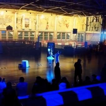 The Coney Island Performance, with a ballerina and a ballerino standing in a cast light in the middle of an open floor, with everything else visible cast in a dark-blue light