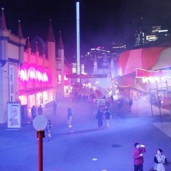 The Main Drag, with a view from above the ground of a walkable street lined with arcades and similar building which is all cast in a magenta-lavender hue aided by smoke machines placed at intervals down the street