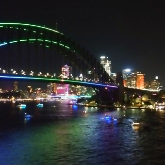 The Ferris Wheel's View, with an aeriel view of Sydney Harbour Bridge trimmed with green and blue lights which is reflected in the water beneath it, with Sydney CBD lit up behind the bridge