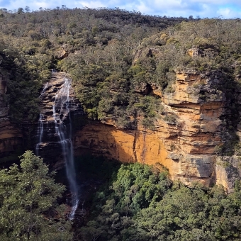 Long Falls, Princes Rock Lookout, Wentworth Falls, with a long waterfall being caught by the wind and wisped into the valley to the right, with surrounding orange cliffs left and right, with green trees above and below