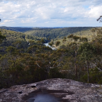 Robertson Knoll Lookout, Engadine Track, with the river barely visible amongst rolling trees and hills, with a rocky cliff in the foreground, with pale-blue clouds above