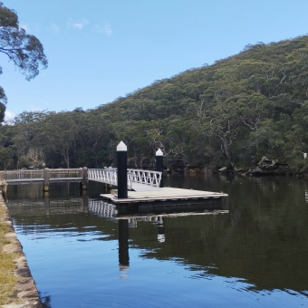 Pool Flat Jetty, Audley, with a small floating weir in the middle of the river which is attached to the mainland by bridge, with dense trees and blue sky reflected in the river