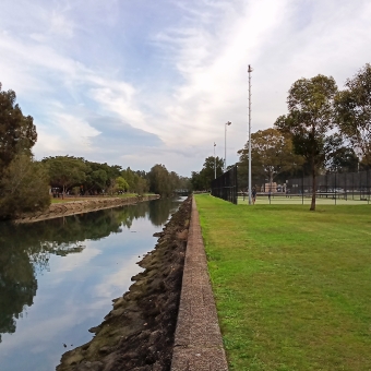 Hawthorne Canal, Haberfield, with shallow canal on the left, with grass then tennis courts on the right, with grey-ish clouds above