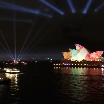 Iconic Sydney, from OPT, with the Sydney Opera House projected with orange and green and red, with two ferries sailing under a fanned-out spread array of spotlights