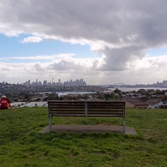 Distant City, Dudley Page Reserve, Dover Heights, with the outline of Sydney CBD and the Harbour Bridge silhouetted in the distance, with a wooden bench above green grass in the foreground